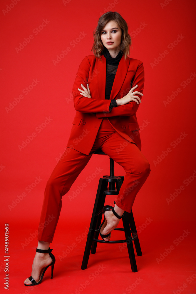 Naklejka premium High fashion photo of a beautiful elegant young woman in a pretty red suit, jacket, pants, trousers, black blouse posing on background. Monochrome, total red. Model sits on chair