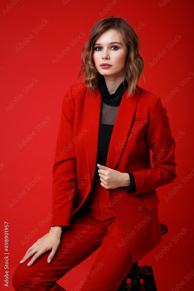 High fashion photo of a beautiful elegant young woman in a pretty red suit, jacket, pants, trousers, black blouse posing on background. Monochrome, total red. Model sits on chair High fashion photo of a beautiful elegant young woman in a pretty red suit, jacket, pants, trousers, black blouse posing on background. Monochrome, total red. Model sits on chair