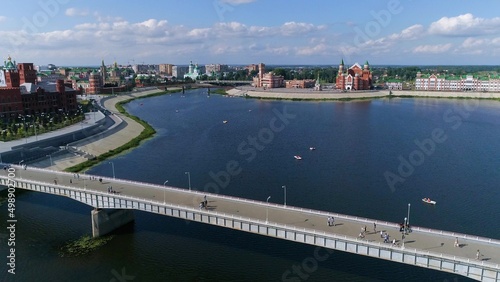 Flying over the river in the city limits. The banks are covered with concrete slabs. A footbridge crosses the river. Boats and catamarans float on water. The concept of a comfortable urban environment