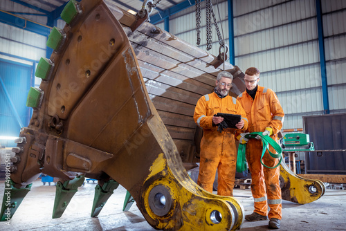Engineer with apprentice next to digger bucket in engineering factory