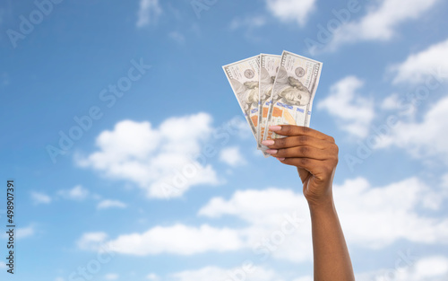 finance, currency and people concept - close up of female hand holding dollar money over blue sky and clouds background