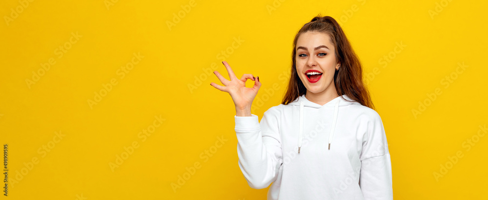 Positive good-looking young brunette woman in white casual style sweatshirt, smiling happy and doing ok sign with hand. Indoor studio shot on yellow background with copy space for text Positive good-looking young brunette woman in white casual style sweatshirt, smiling happy and doing ok sign with hand. Indoor studio shot on yellow background with copy space for text