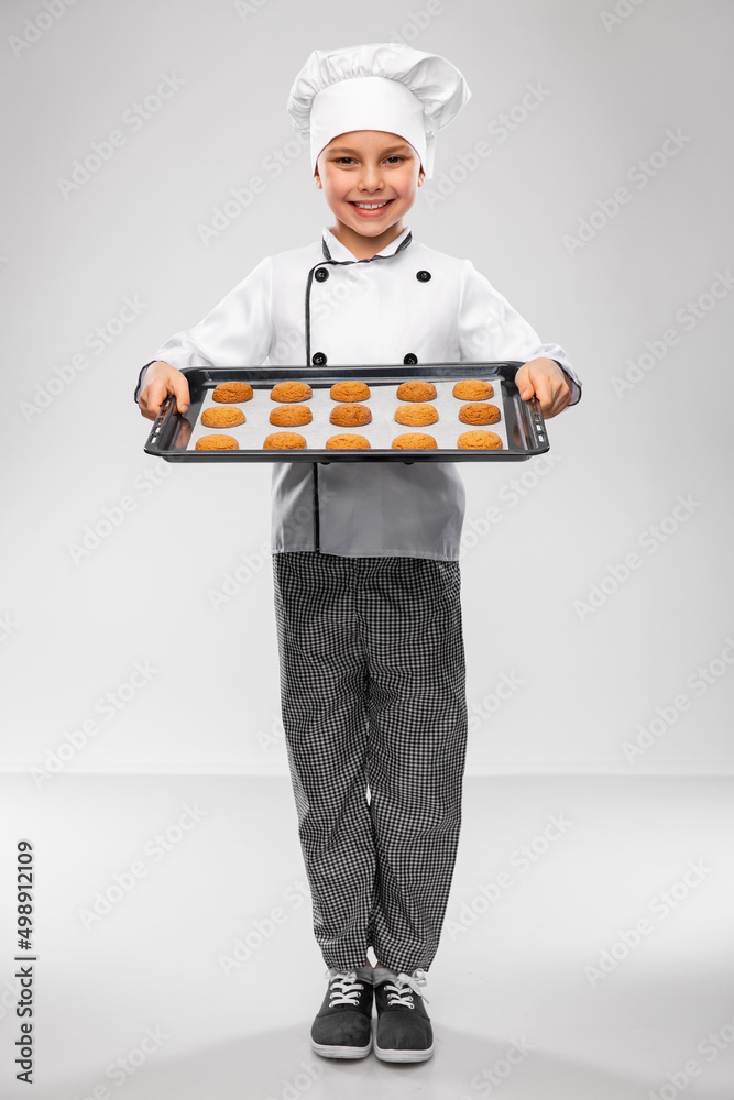 cooking, culinary and profession concept - happy smiling little boy in chef's toque and jacket holding baking tray with oatmeal cookies over grey background cooking, culinary and profession concept - happy smiling little boy in chef's toque and jacket holding baking tray with oatmeal cookies over grey background