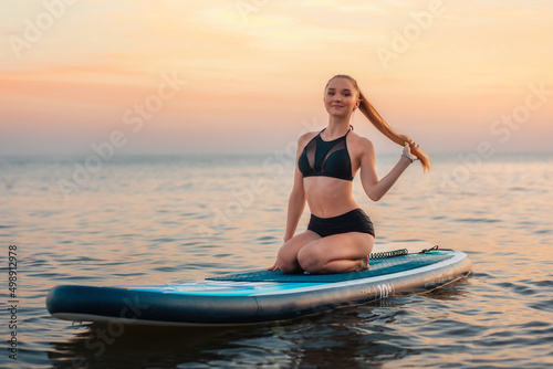 Surfing. Pretty young slender woman sitting on a sup board. In the background, the ocean and the sunset. Summer activity © _KUBE_