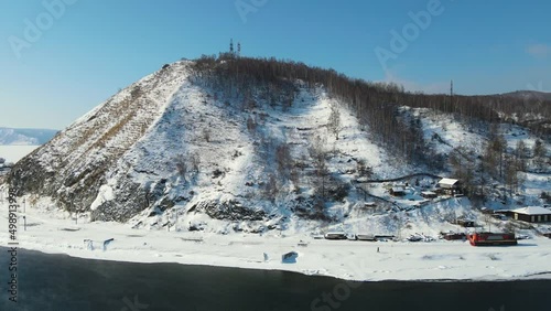 Drone movies around a wooded hill. The mountain is skirted by a railway. On the shore of the non-freezing lake at the station there is a red locomotive. Northern village in the wilderness on winter.