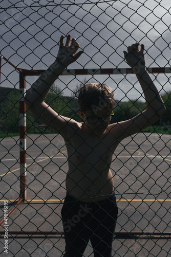 BOY WITH GLASSES AND NO SHIRT IS RESTING LEANING ON THE FENCE OF A SOCCER FIELD. BEHIND IS A RED AND WHITE GOAL.