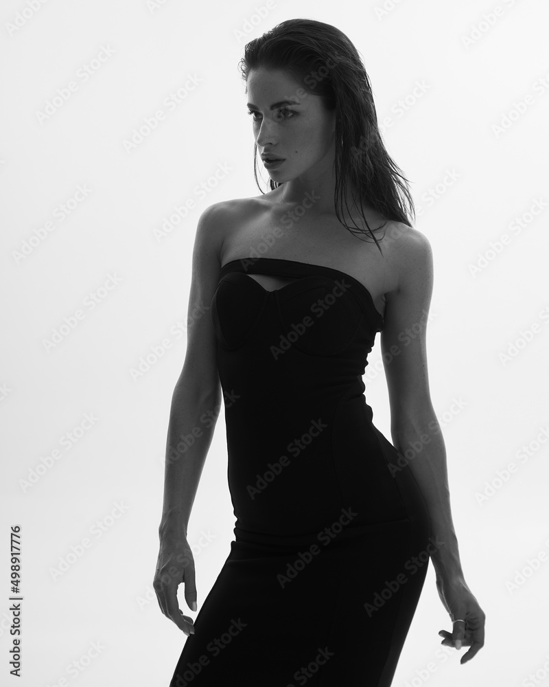Fototapeta premium Portrait of young attractive woman model with brown long curly hair, black dress, long legs with tan skin, slim body. The concept of a beautiful elegant figure, body positive, healthy lifestyle.