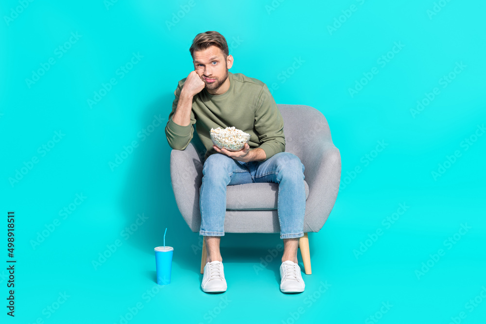 Photo of bored man hold popcorn soda sit armchair watch movie wear pullover isolated blue color background Photo of bored man hold popcorn soda sit armchair watch movie wear pullover isolated blue color background