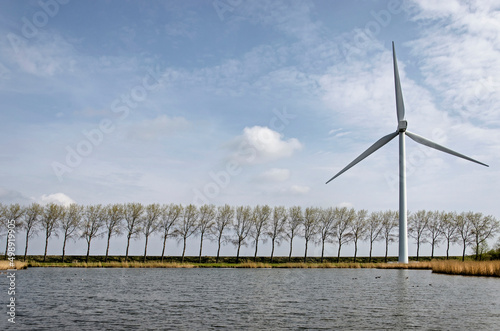 Middelharnis, The Netherlands, March 30, 2022: view across a lake towards a dike with a wind turbine and a row of trees all bend in the same direction by the prevailing westerly winds photo