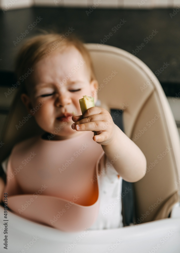 10 months old baby sitting in high chair, with a silicone bib, eating avocado, grimacing. 10 months old baby sitting in high chair, with a silicone bib, eating avocado, grimacing.