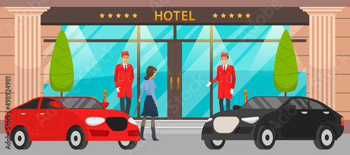 Doormen in red uniform next to door inviting to hotel. Girl with suitcase near entrance to building. Main front entrance to luxury hotel and premium cars. Staff and visitors near large building