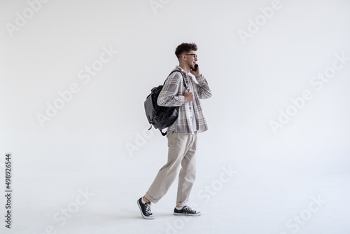 Full length portrait of a smiling young male student dressed in plaid shirt with a backpack talking on mobile phone isolated over white background