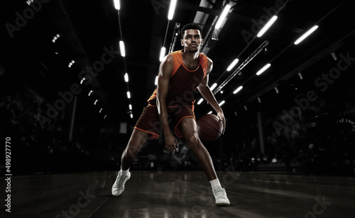 African-american young basketball player in action and motion in flashlights over dark gym background. Concept of sport, energy and dynamic, healthy lifestyle. Arena's drawned