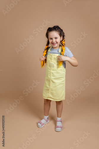 Delighted little girl holding yellow kanekalon pigtails with hands, looking at camera with smile on face indulging wearing yellow jumpsuit and gray t-shirt on beige background.