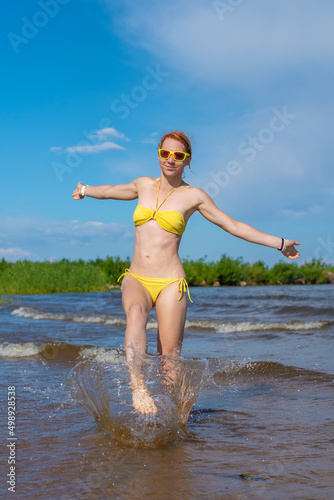 Portrait of a beautiful young sexy caucasian woman with wet red hair playing with water on the beach in a yellow bikini and yellow sunglasses. Kallaste beach, Lake Peipus, Estonia.
