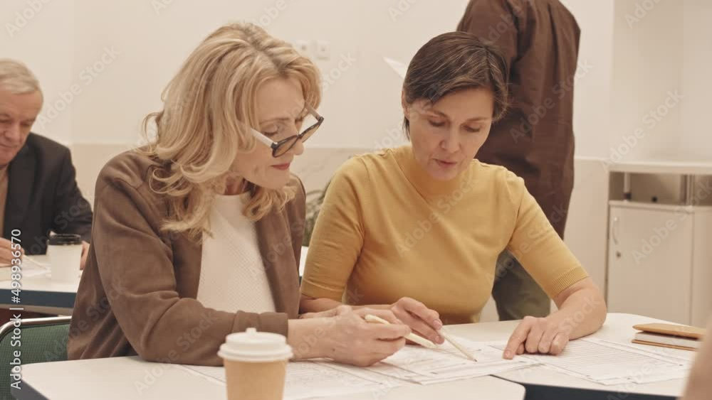 Waist up slowmo of two Caucasian mature women discussing Math problem while looking at paper practice test, sitting together at desk in classroom