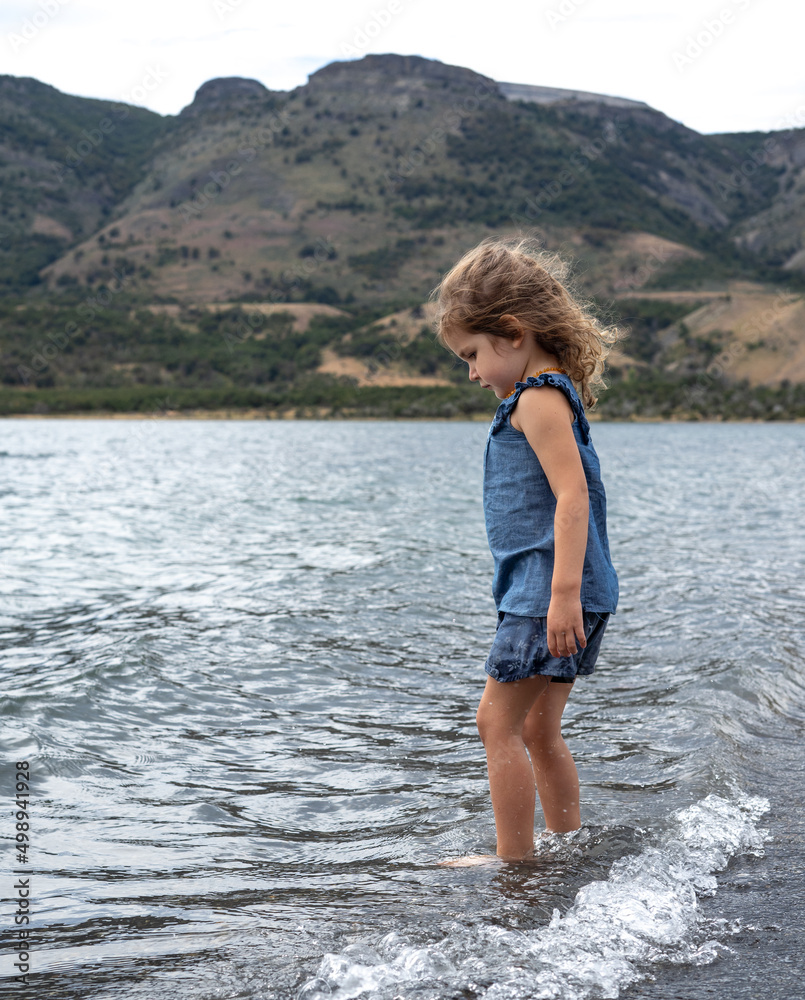 Little pretty girl in blue dress staying in water of the lake near mountains. High quality photo