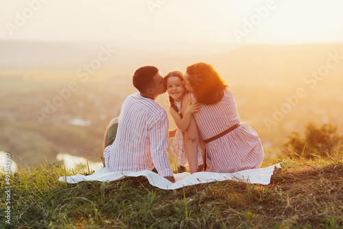 Young beautiful family with a little daughter on the hill at sunset. Parents hug and kiss their happy daughter. Family with a small child in nature.