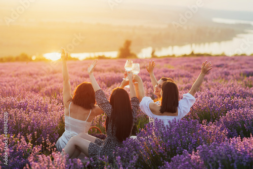 Group of young friends having picnic in the lavender field, toasting with wine at sunset. Happy friends sitting together at nature. Holiday and summer vacation concept.