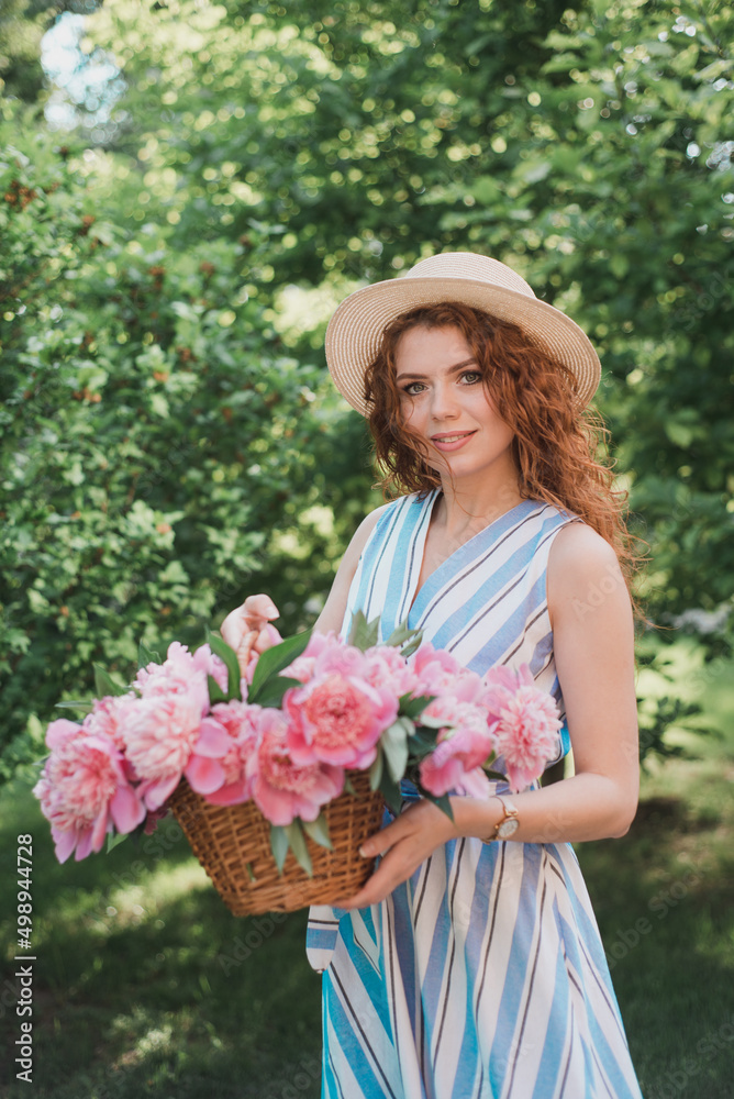 Fototapeta premium Portrait of young redhead curly woman in straw hat and linen stripe dress with a basket and a pink peonies bouquet in the garden