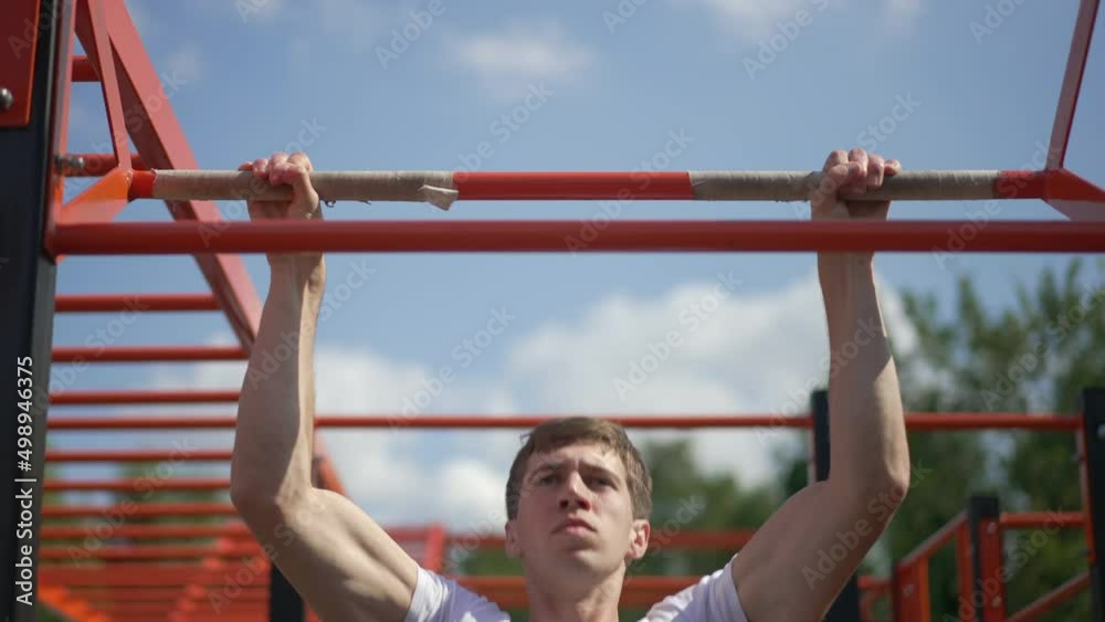 Front view serious sportsman doing pull ups on gymnastics bar in sunlight outdoors. Portrait of Caucasian fit man working out on sunny day in spring summer park in slow motion Front view serious sportsman doing pull ups on gymnastics bar in sunlight outdoors. Portrait of Caucasian fit man working out on sunny day in spring summer park in slow motion