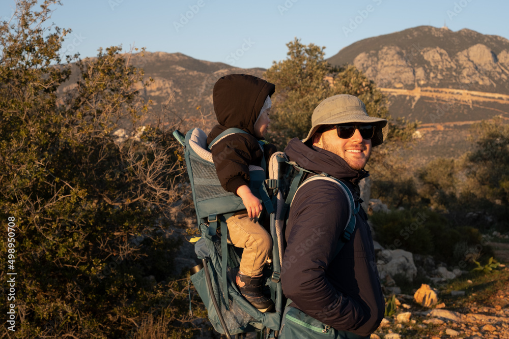 Man wearing sunhat, sunglasses carrying baby,toddler in backpack.Young handsome father traveling,hiking with kid, child of 1,2 years old.Family trip in nature,mountains,cold weather near sea