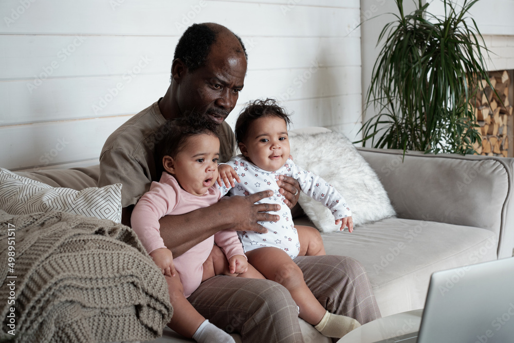Portrait of mature African American father sitting on sofa in living room with twin baby daughters on his lap watching cartoons in Internet