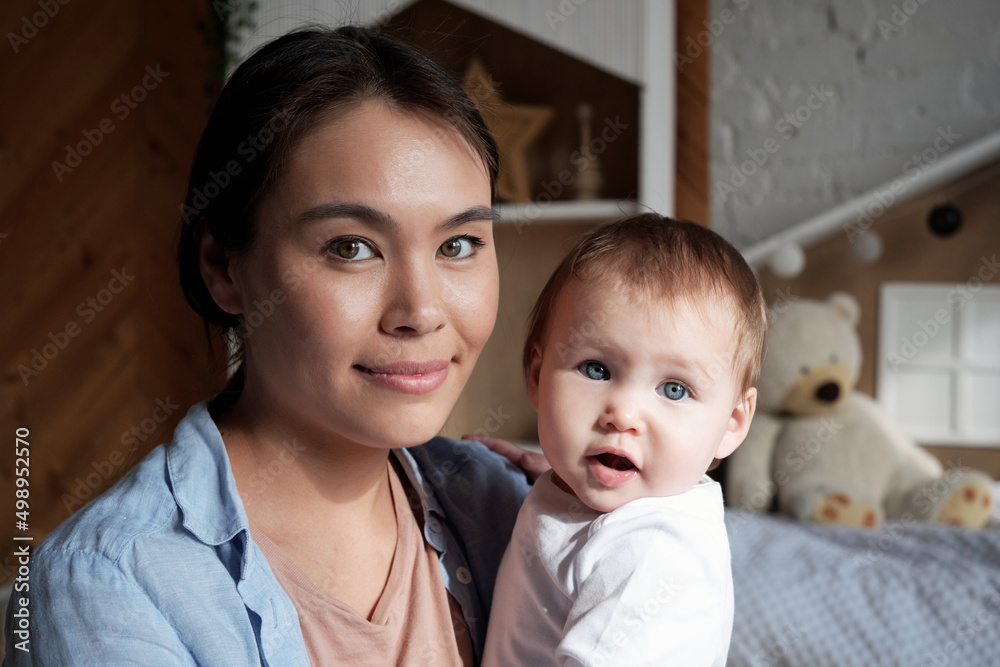 Close-up portrait of beautiful young mother spending time at home holding baby daughter in arms looking at camera Close-up portrait of beautiful young mother spending time at home holding baby daughter in arms looking at camera