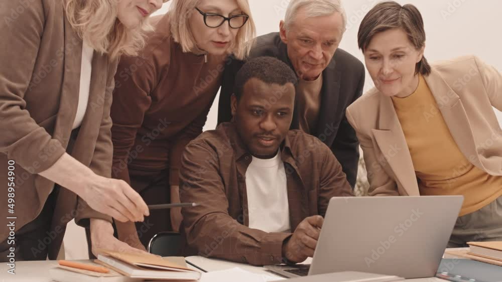 Slowmo of male Black IT teacher showing and explaining how to use laptop computer to group of adult students standing around his desk and pointing on laptop screen asking questions