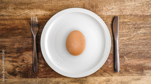 Raw egg on white plate against wooden table with knife and fork, top view
