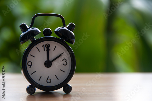 Black alarm clock isolated on blurred nature background. The clock set at 2 o'clock. Copy space concept