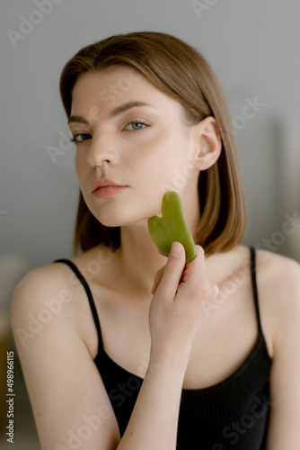 Young woman with a beautiful hairstyle with a black tank top makes a massage with a green quartz stone gua sha on her face. Looks at the camera in the bedroom.