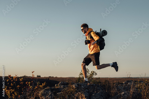 Travel Man Dressed in Windbreaker with Backpack and Camping Mat, Jumping on the Top of Hill During Sunset, Male Nomad with Digital Camera Enjoy His Best Solo Trip
