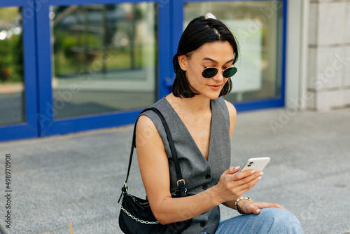 Stylish short-haired girl in dark sunglasses with bag smiles outdoors. Brunette woman in grey vest is holding phone and listening to music outside