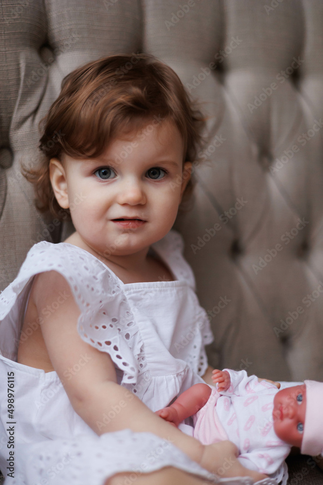 Little girl portrait. Small baby kid playing with a doll. Serious little daughter looking attentively and thoughtfully. One-year-old girl in whire dress sitting on beige sofa in big room. Sweet kid Little girl portrait. Small baby kid playing with a doll. Serious little daughter looking attentively and thoughtfully. One-year-old girl in whire dress sitting on beige sofa in big room. Sweet kid