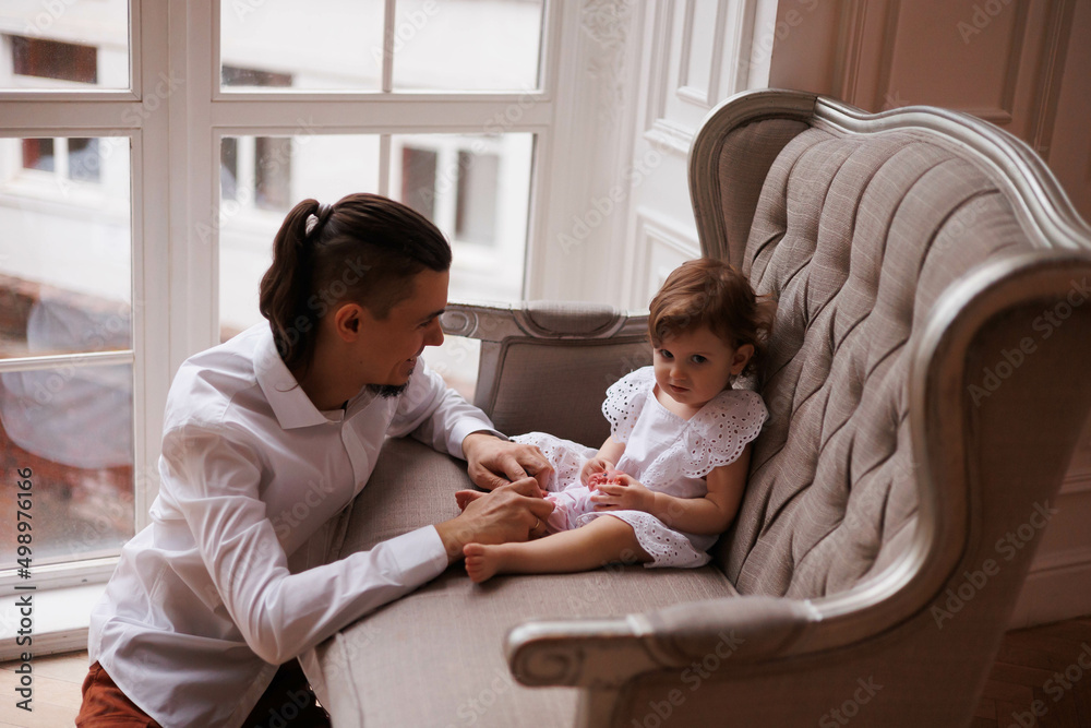 Handsome happy father playing with legs of little daughter on beige sofa indoors. Young man in white shirt with his 1-year-old daughter in white dress, holding her legs. family tenderness Handsome happy father playing with legs of little daughter on beige sofa indoors. Young man in white shirt with his 1-year-old daughter in white dress, holding her legs. family tenderness