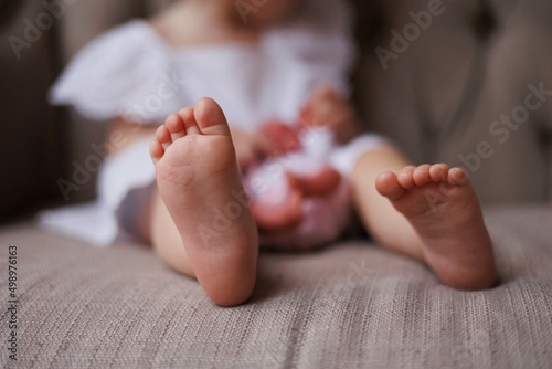 Closeup of little baby girl's feet. Daughter sitting on sofa barefeet. Sweet girl in white dress. Love and tenderness