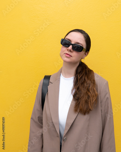 Young caucasian millennial female in sunglasses and brown blazer on a yellow background