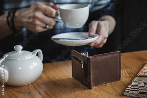 Close-up of a brown leather wallet on a wooden table against the background of a man drinking tea from a glass. Horizontal orientation, no face, focus in the foreground