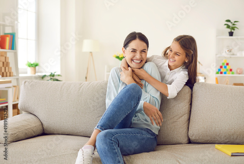 Happy mother hugging together and having fun with her teenage daughter at home on weekend. Cheerful child tickles his positive young mother sitting on sofa in living room. Family concept. © Studio Romantic