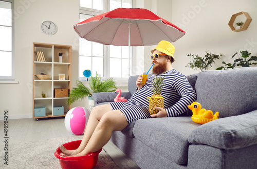 Funny man in living room at home imagines that he is resting on sea and sunbathing on beach. Chubby man wets his feet in plastic bowl and drinks cocktail while sitting on sofa under beach umbrella. photo