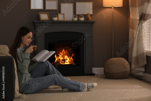 Beautiful young woman with cup of coffee reading book on floor near fireplace at home. Space for text
