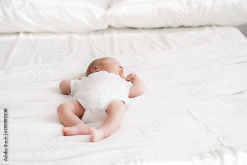 funny newborn baby in white bodysuit sleep sweetly on large white bed