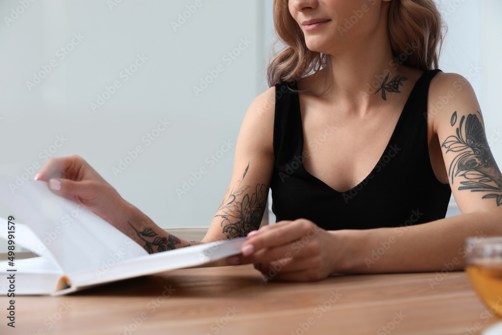 Beautiful woman with tattoos on body reading book at table indoors, closeup Beautiful woman with tattoos on body reading book at table indoors, closeup