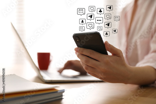 Different virtual icons and young woman using smartphone and laptop at table indoors, closeup. SMM (Social media marketing) concept photo