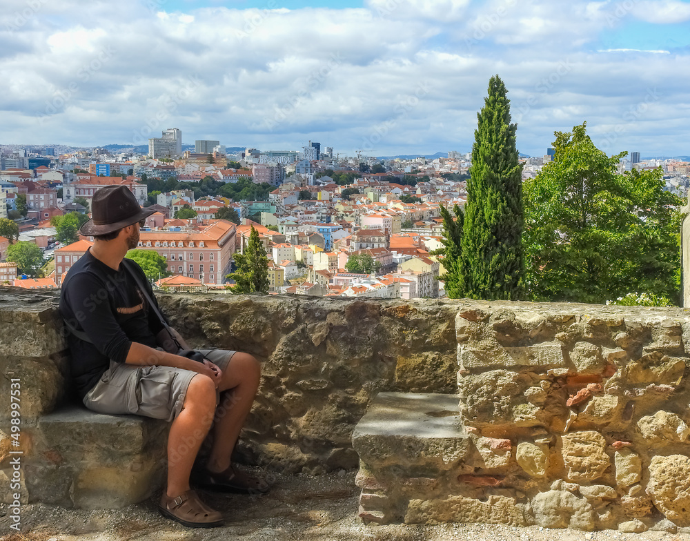 Fototapeta premium Lisbon, Portugal - July 07 2019: Castelo de Sao Jorge: caucasian man seated on the walls of the castle. He wears a hat, a black shirt and brown bermudas