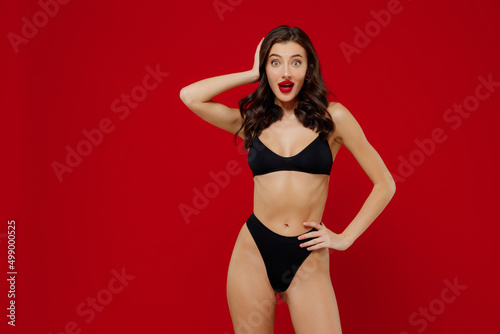 Young shocked sexy brunette woman 20s with perfect fit body wear black underwear look camera with opened mouth hold head isolated on plain red background studio portrait. People female beauty concept