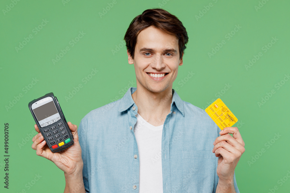 Excited jubilant young brunet man 20s years old wears blue shirt hold wireless modern bank payment terminal to process acquire credit card payments isolated on plain green background studio portrait. Excited jubilant young brunet man 20s years old wears blue shirt hold wireless modern bank payment terminal to process acquire credit card payments isolated on plain green background studio portrait.