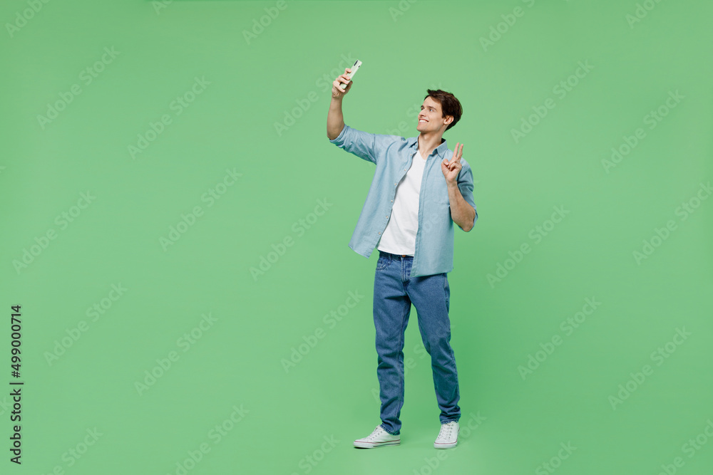 Full size side view fun young brunet man 20s wear blue shirt get video call use mobile cell phone conducting pleasant conversation show victory sign isolated on plain green background studio portrait Full size side view fun young brunet man 20s wear blue shirt get video call use mobile cell phone conducting pleasant conversation show victory sign isolated on plain green background studio portrait