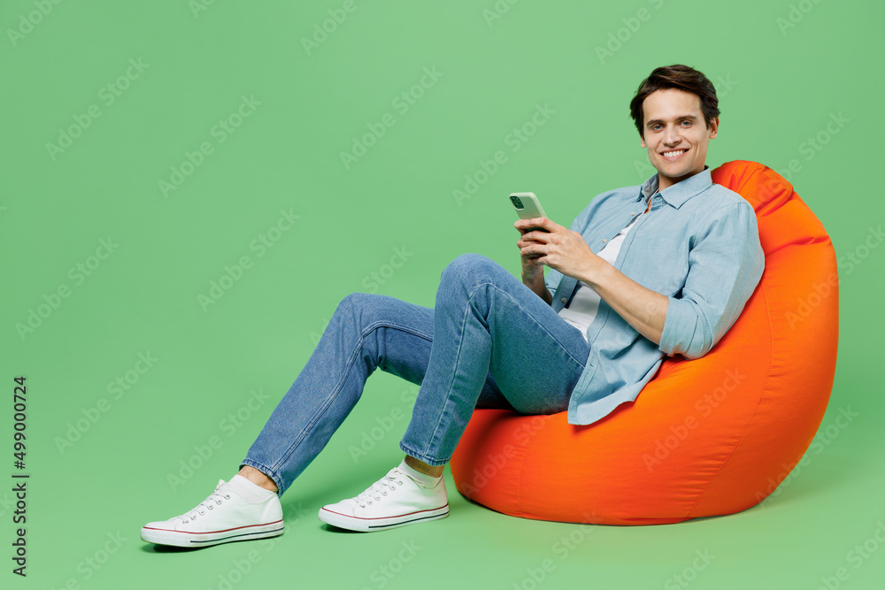 Full size young brunet man 20s wears blue shirt sit in bag chair hold in hand use mobile cell phone typing browsing chatting send sms looking camera isolated on plain green background studio portrait. Full size young brunet man 20s wears blue shirt sit in bag chair hold in hand use mobile cell phone typing browsing chatting send sms looking camera isolated on plain green background studio portrait.